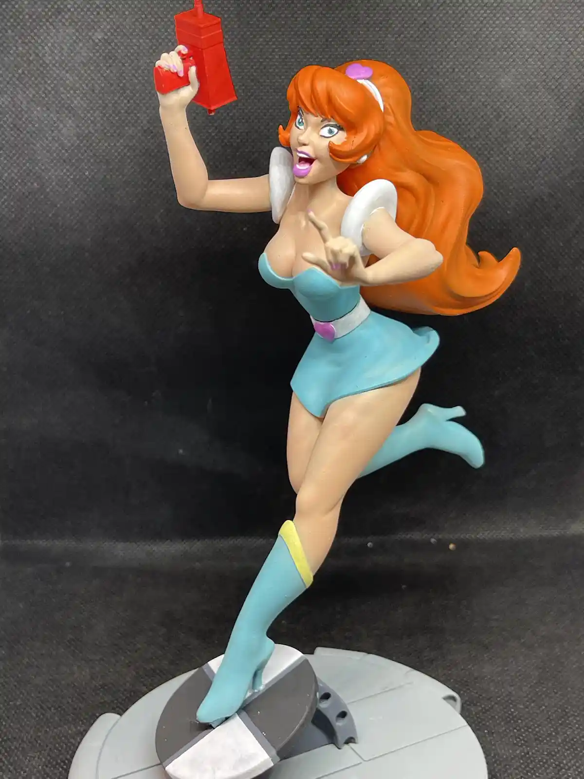 A figurine of a cartoon-style female character with orange hair, wearing a blue outfit and white wings, holds a red object…