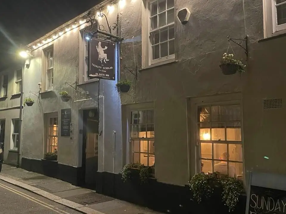 A nighttime photo of a pub named 'Thirsty Scholar' with a sign showing a person riding a horse. Inside one window, a person…