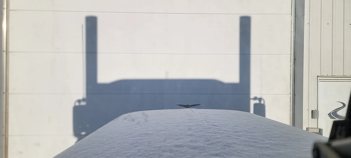 A snow-covered vehicle roof is shown with the shadow of a truck cast upon it, and a small bird perched on the snow. The…