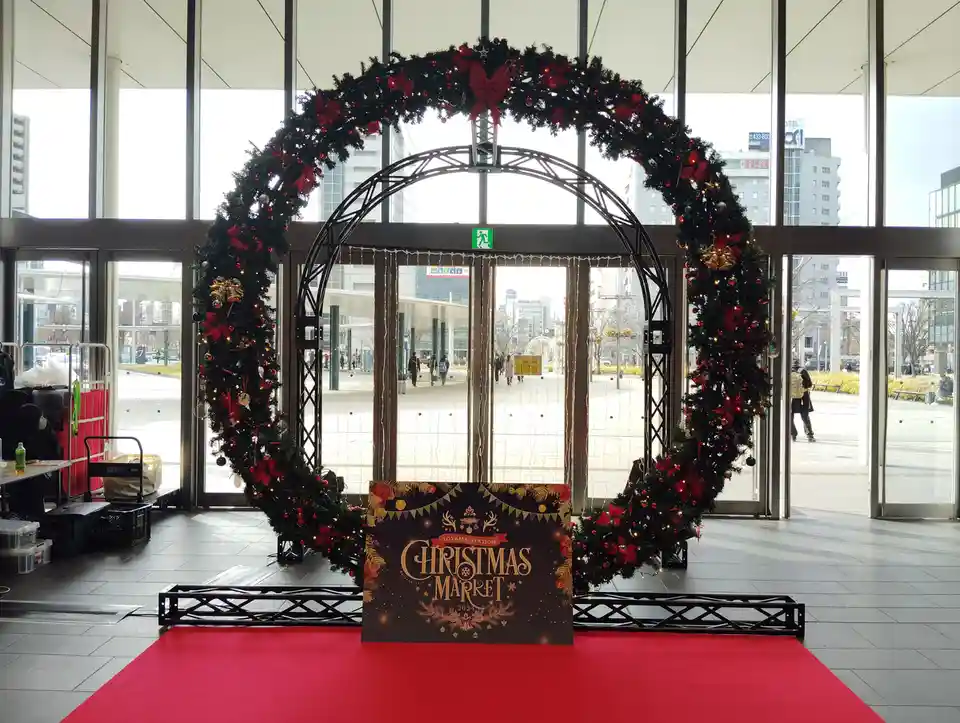 A festive Christmas market entrance at Toyama Station features a large, decorated archway with red bows, lights, and…