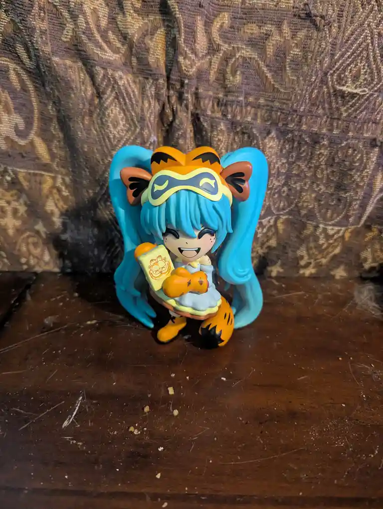 A chibi-style figurine of Hatsune Miku with blue pigtails, wearing an orange tiger-themed mask and outfit, holding a…