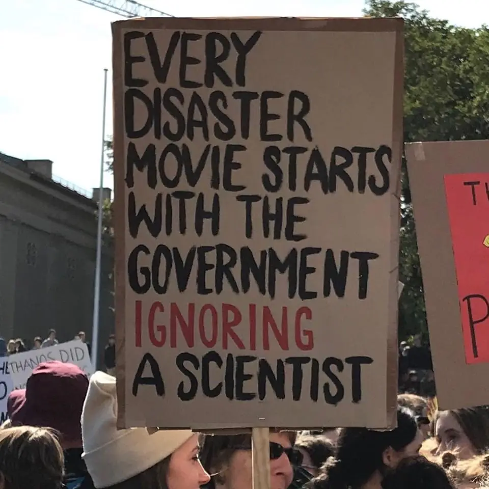 A handmade cardboard protest sign held at an outdoor rally, featuring bold black handwritten text that reads 'EVERY DISASTER…
