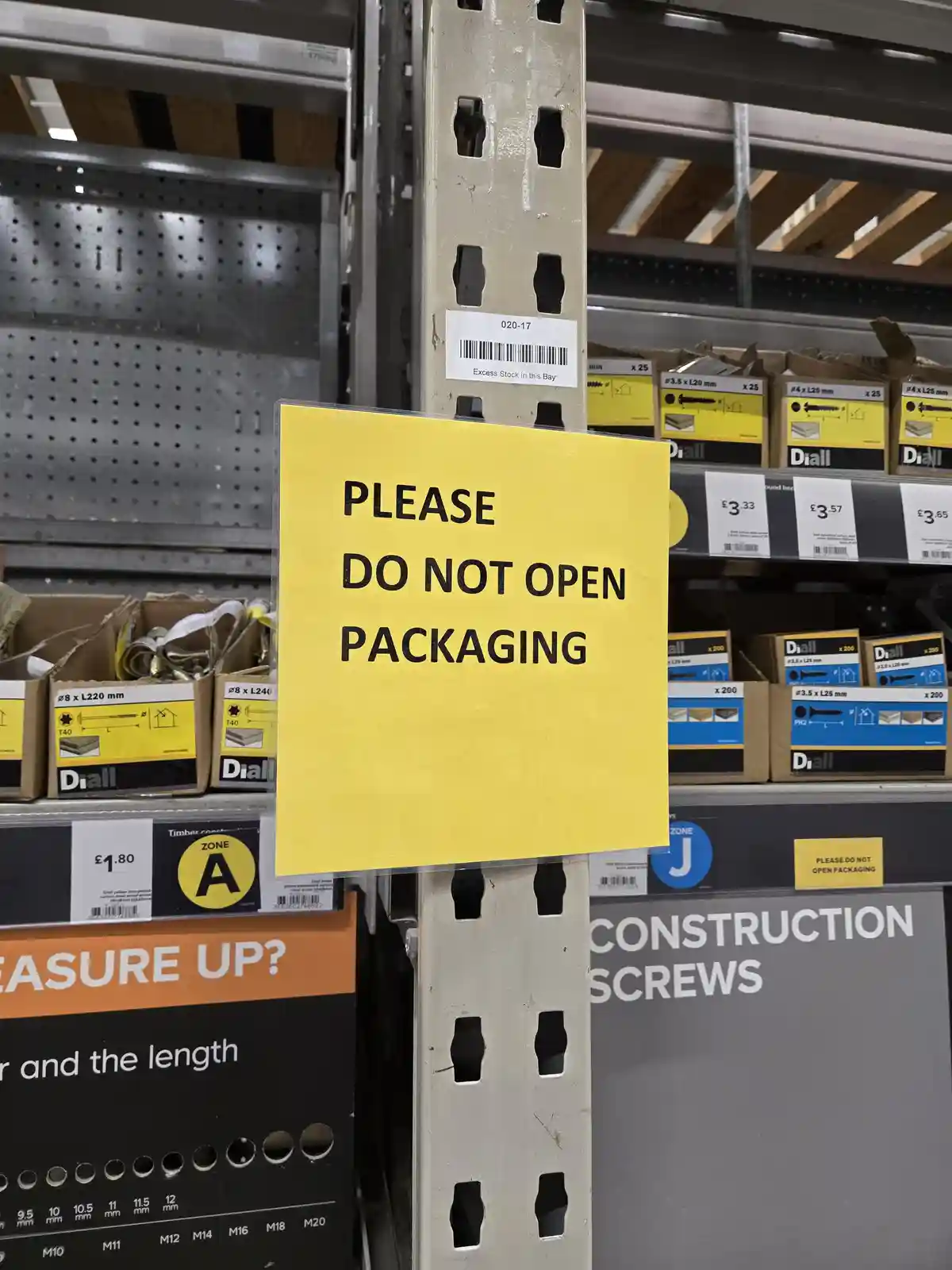 A bright yellow sign in a hardware store aisle reads 'PLEASE DO NOT OPEN PACKAGING' in bold black letters. Behind it…