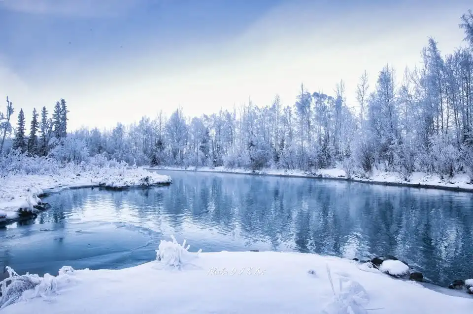 A serene winter landscape featuring a calm river partially frozen, surrounded by snow-covered trees and banks under a pale…