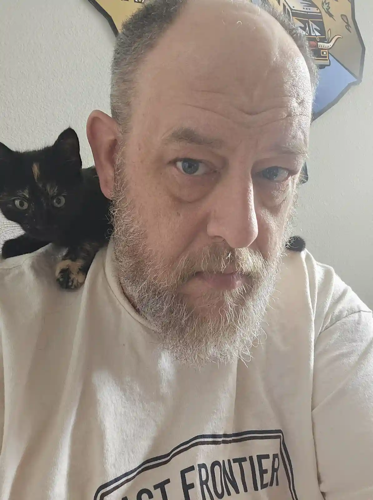 A close-up selfie of a middle-aged man with a graying beard and intense blue eyes, wearing a white sweatshirt with 'LAST…