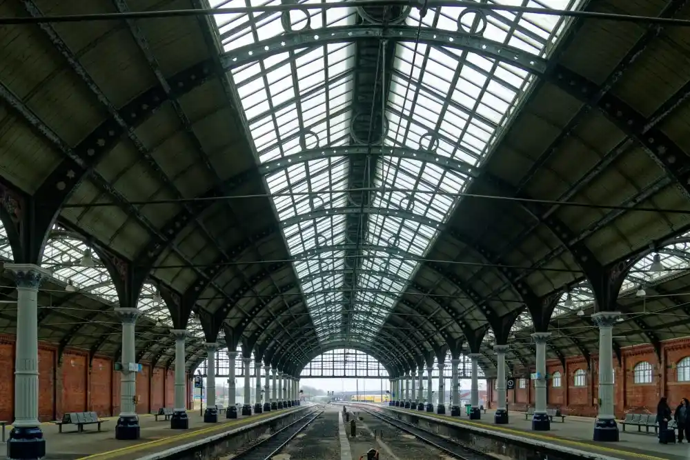 An empty, grand Victorian-era train station with arched glass roof, brick walls, and platforms flanked by pillars and…