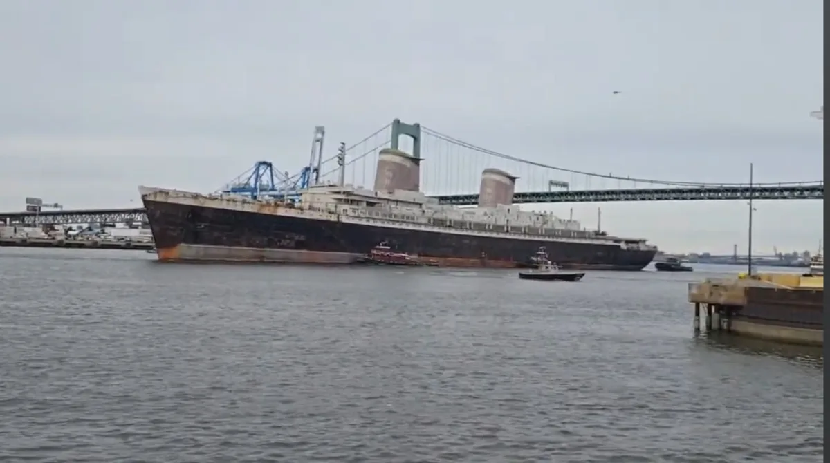 A large, rusty, and visibly deteriorated ship is shown floating in a harbor, with a suspension bridge in the background…
