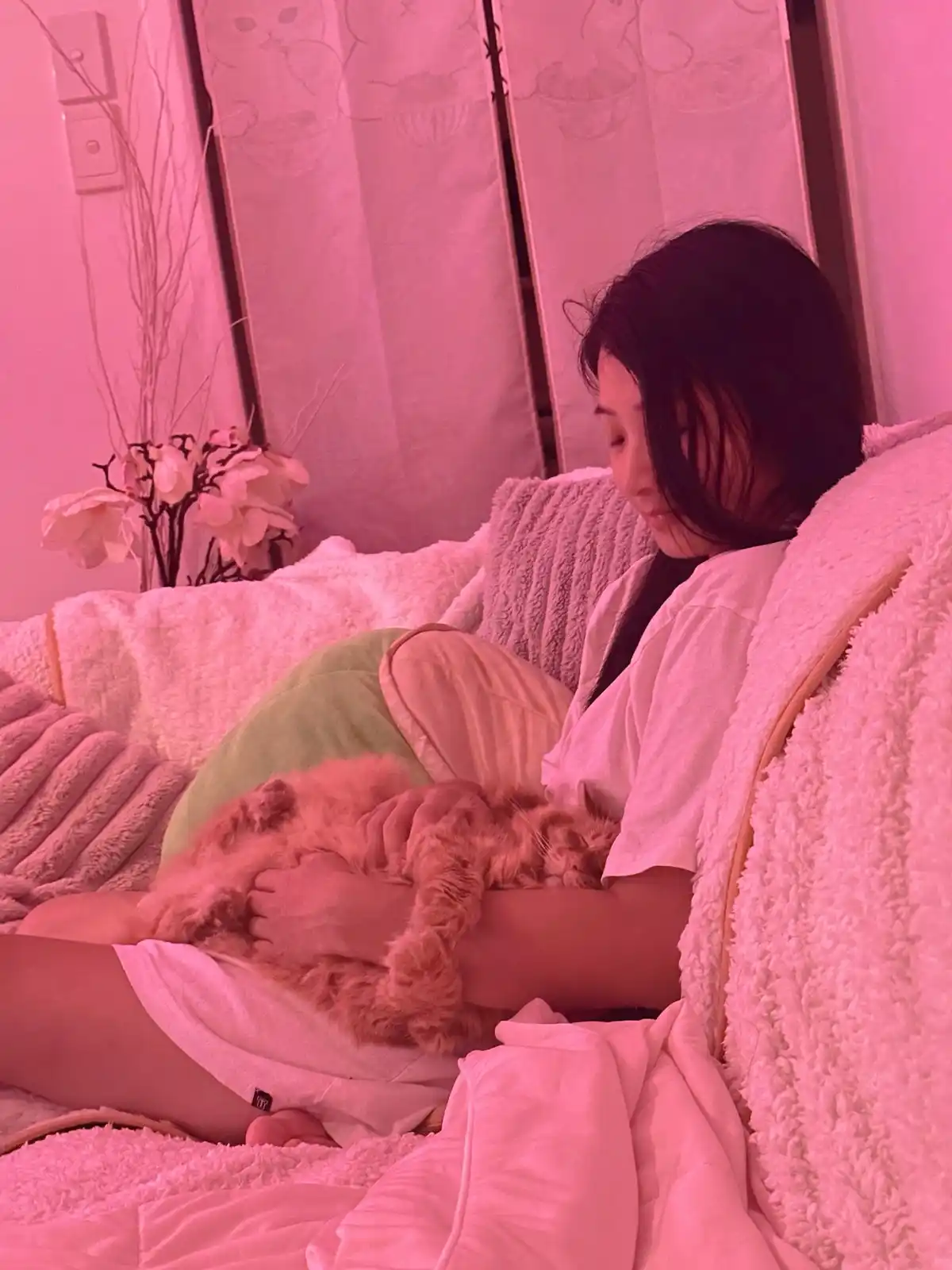 A person with dark hair sits on a couch wrapped in fluffy pink blankets, gently cradling a sleeping orange cat. The entire…