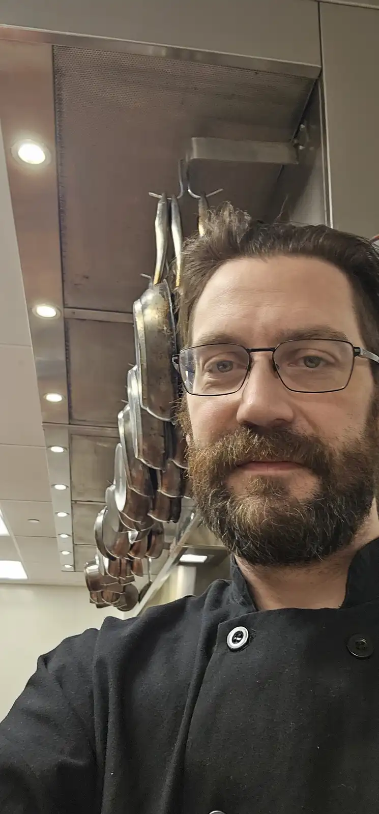 A man in a black chef's coat and glasses takes a selfie in a commercial kitchen, with a large rack of hanging, well-used…