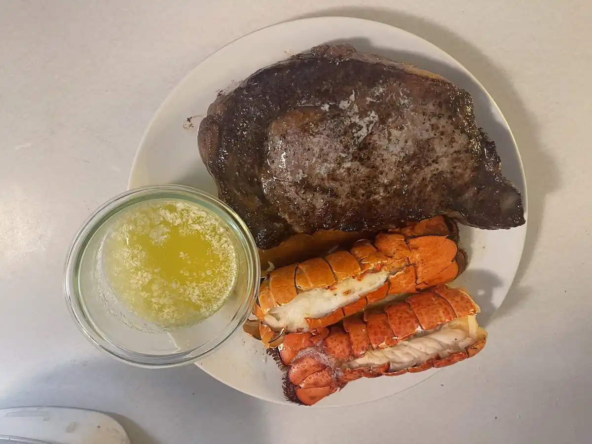 A white plate holds a large, seared steak, two cooked lobster tails with orange shells and white meat, and a small glass…