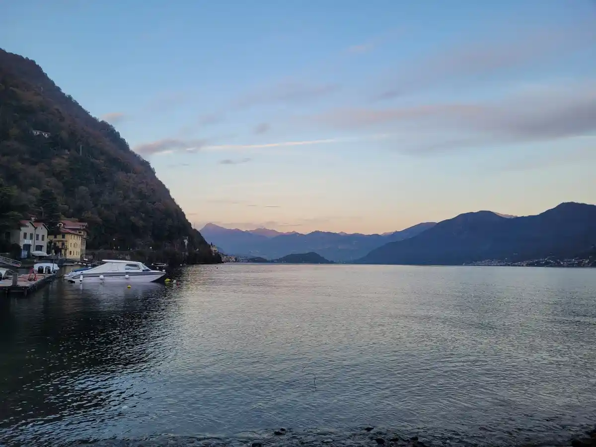 A serene lakeside scene at dusk with calm water, distant mountains, and buildings along the shore; no text is overlaid on…