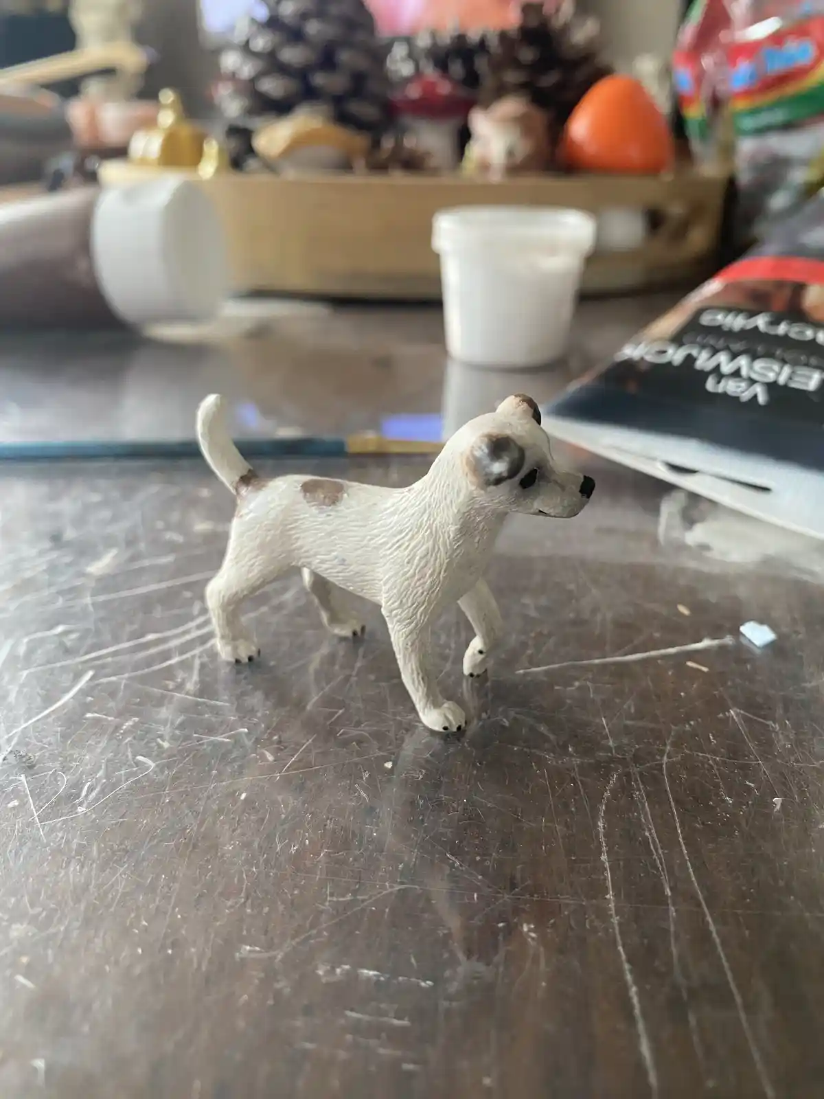 A small, white plastic dog figurine with brown spots stands on a scratched wooden table. The figurine has a slightly worn…