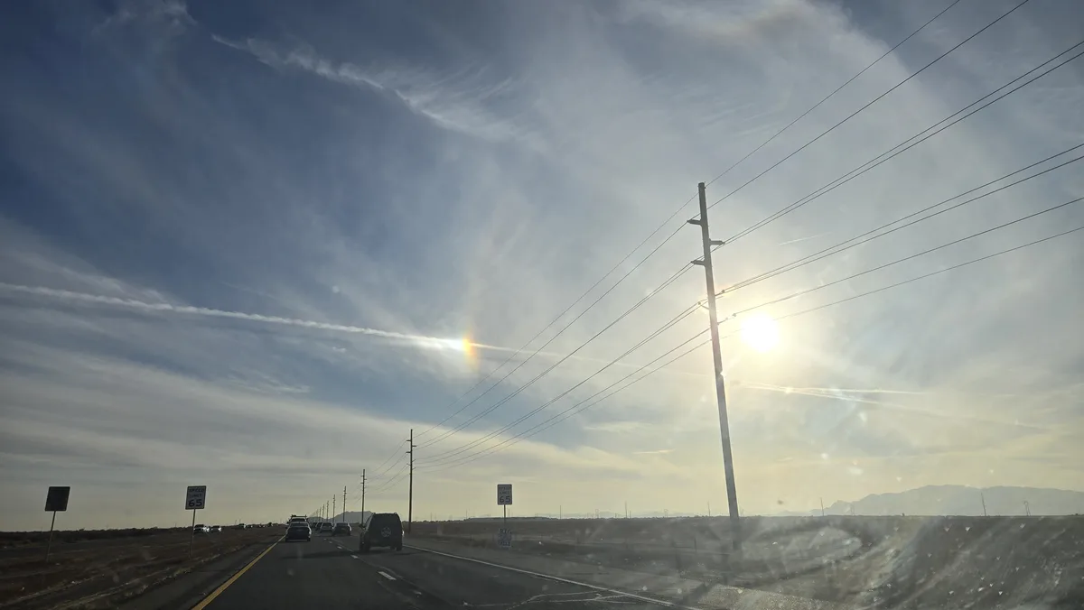 A photo taken from inside a car on a highway shows a bright sun, power lines, and distant mountains under a partly cloudy…