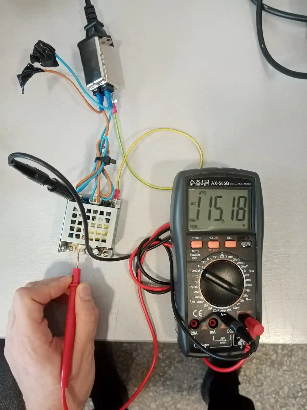 A hand holds a red multimeter probe touching a circuit board connected to a power supply and a digital multimeter. The…