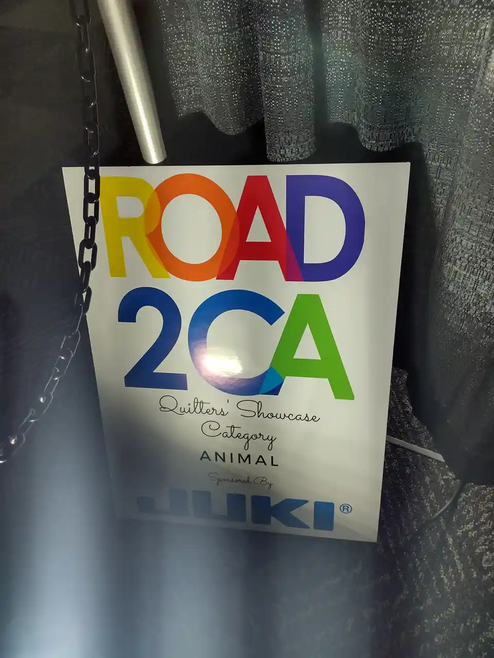 A white sign with colorful, rainbow-gradient text reading 'ROAD 2CA' hangs by a black chain. Below, it says 'Quilters'…