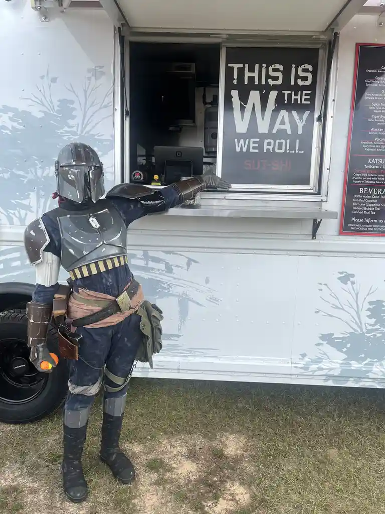 A person in full Mandalorian armor stands at the service window of a food truck, their gloved hand resting on the counter…