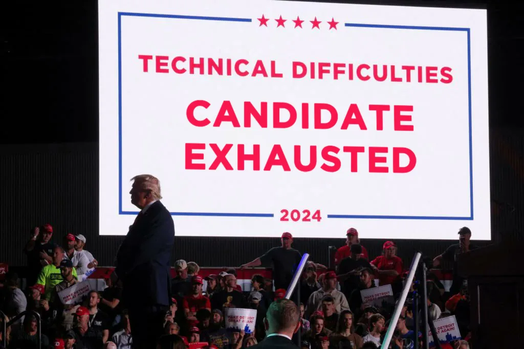 A man in a dark suit stands on a stage in front of a large audience, with a large screen behind him displaying the text…