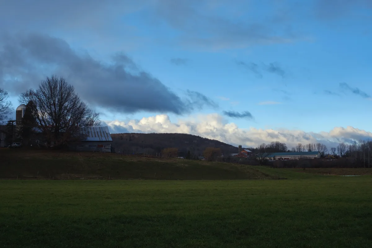 A serene rural landscape featuring a green field in the foreground, rolling hills, scattered farm buildings including a silo…