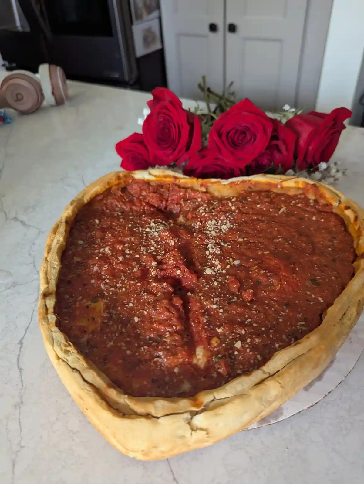 A heart-shaped deep-dish pizza with thick tomato sauce and grated cheese sits on a marble countertop, with a bouquet of red…