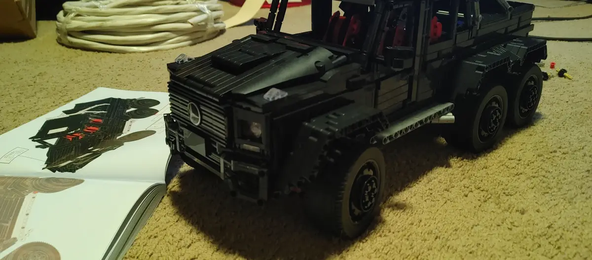 A large, black LEGO Technic Mercedes-Benz G-Wagen model sits on a beige carpet next to an open instruction manual showing a…