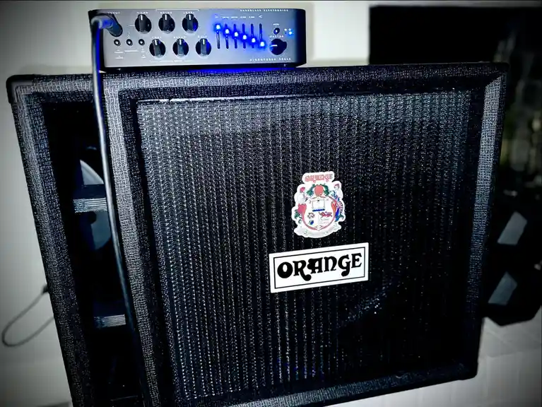 A close-up photo of a black Orange brand guitar amplifier with a textured grille cloth, featuring the Orange logo and crest…