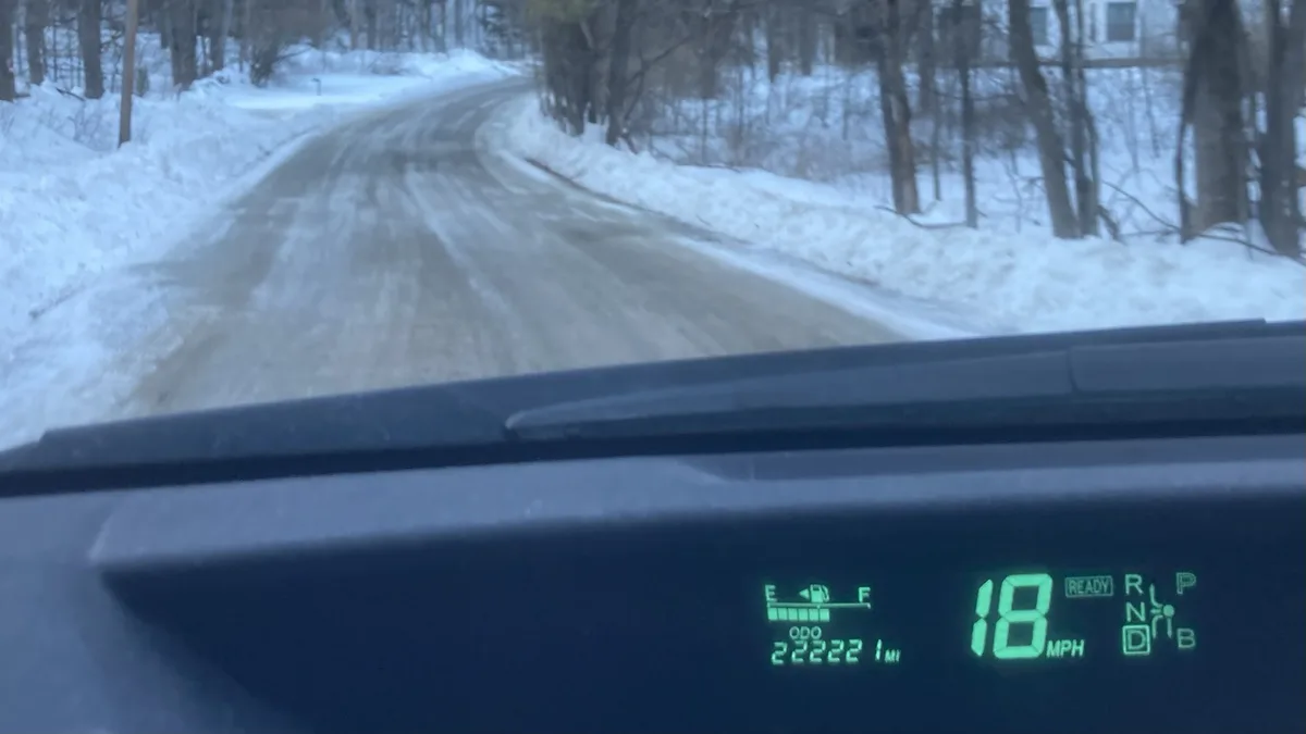 A car's dashboard is visible from inside a vehicle driving on a snowy, winding road through a wooded area. The digital…