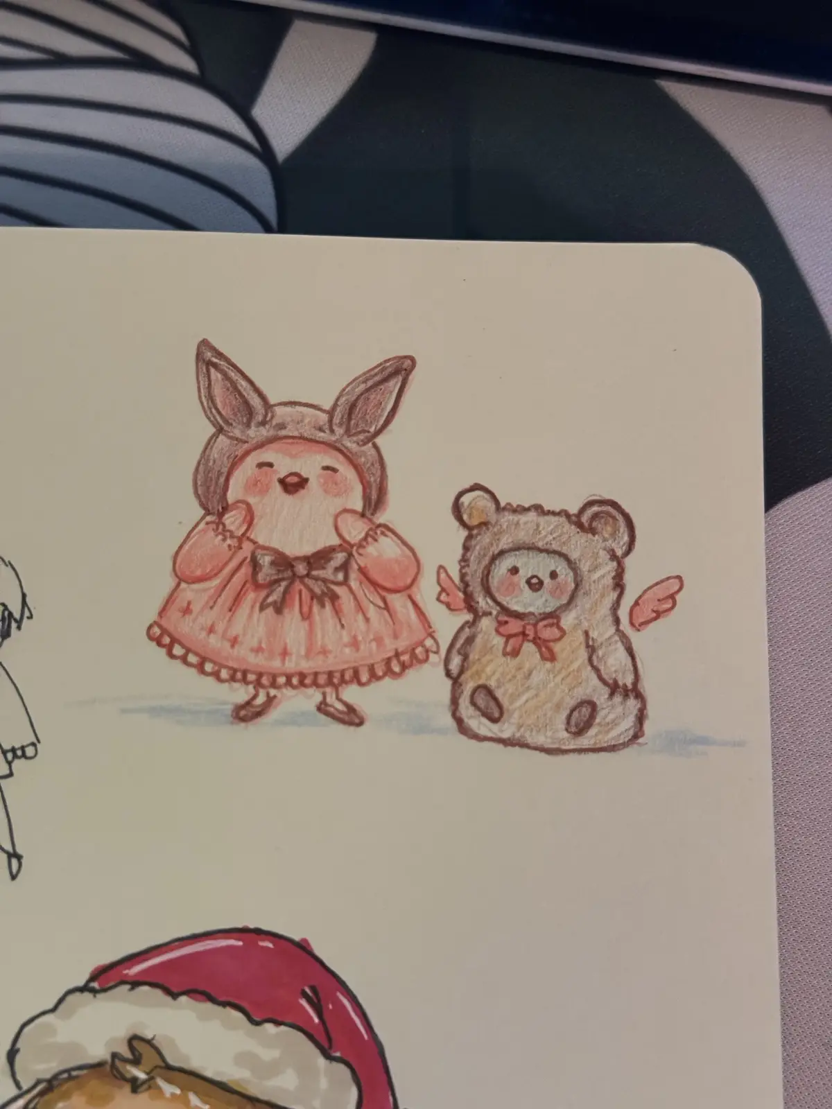 A hand-drawn illustration on paper shows two cute, chibi-style animal characters: one wearing a pink dress with bunny ears…