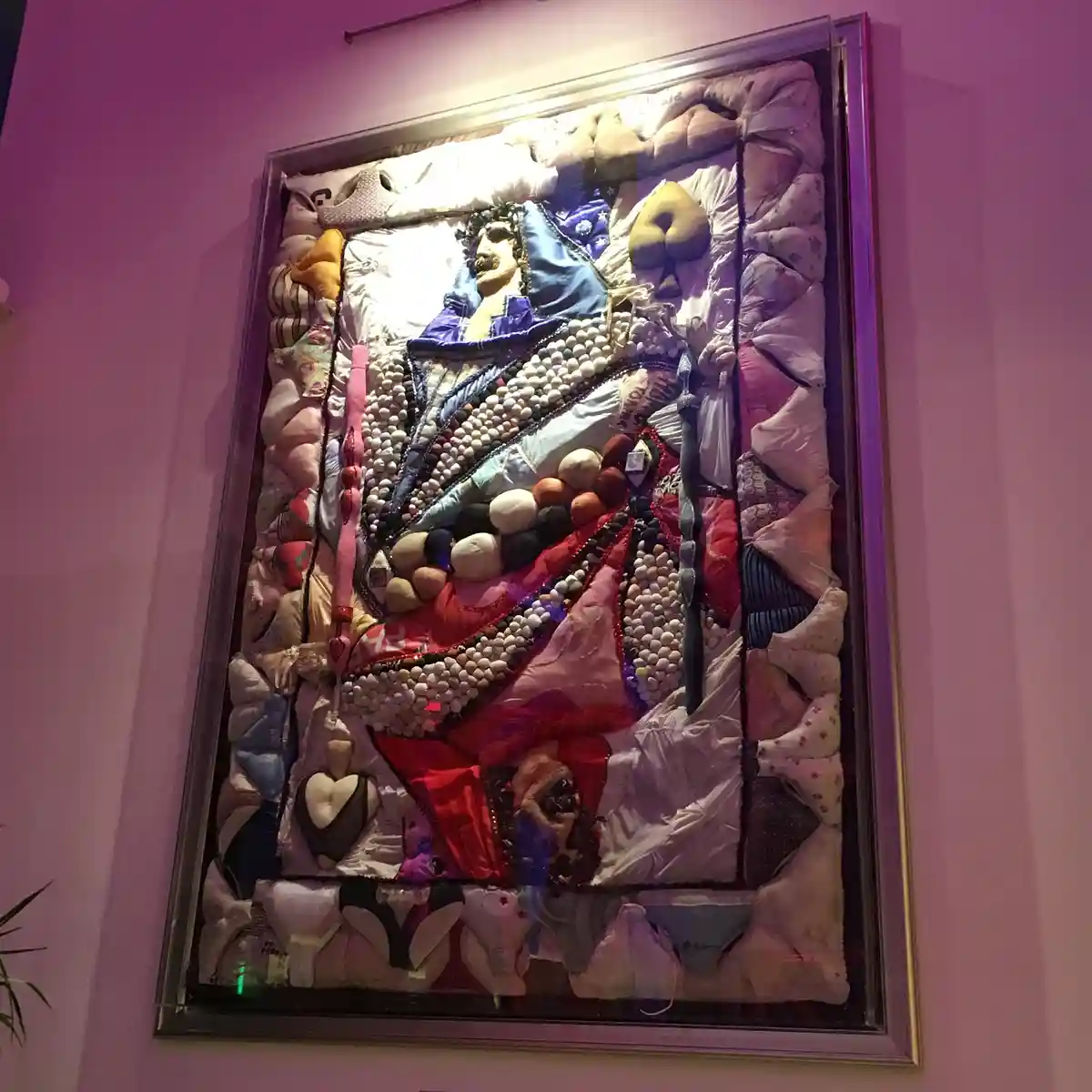 A framed, three-dimensional artwork depicts a stylized queen figure made from various fabrics, beads, and plush items…