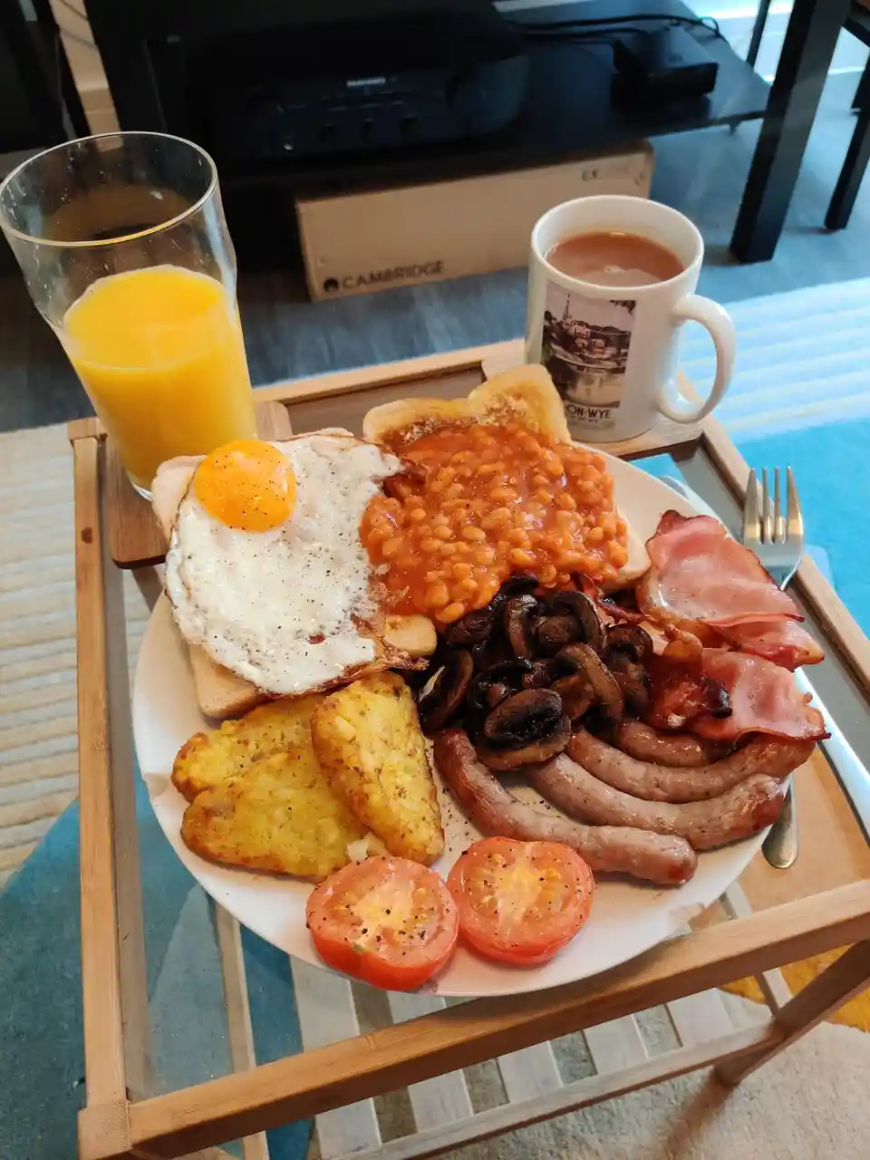A plate of full English breakfast with fried egg, baked beans, sausages, bacon, mushrooms, hash browns, and tomato slices…