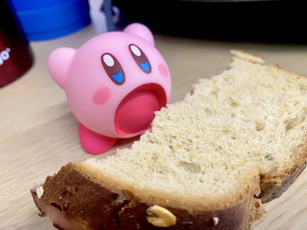 A pink Kirby figurine with wide, surprised eyes and an open mouth sits next to a slice of whole grain bread on a wooden…