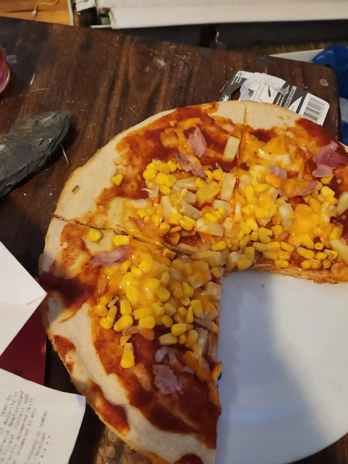 A partially eaten pizza with ham, pineapple, and corn toppings sits on a wooden table next to a white plate. The overlaid…