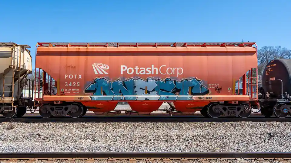 A side view of an orange PotashCorp railcar with the company logo and number POTX 3425, featuring large blue graffiti art…