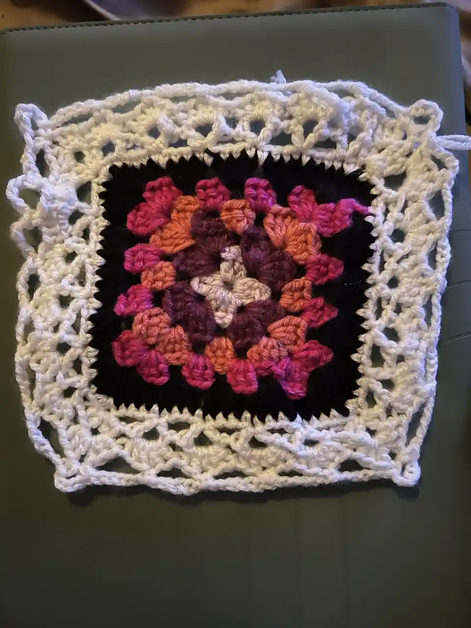 A handmade crocheted square with a white lace border and a colorful floral pattern in the center, resting on a dark green…