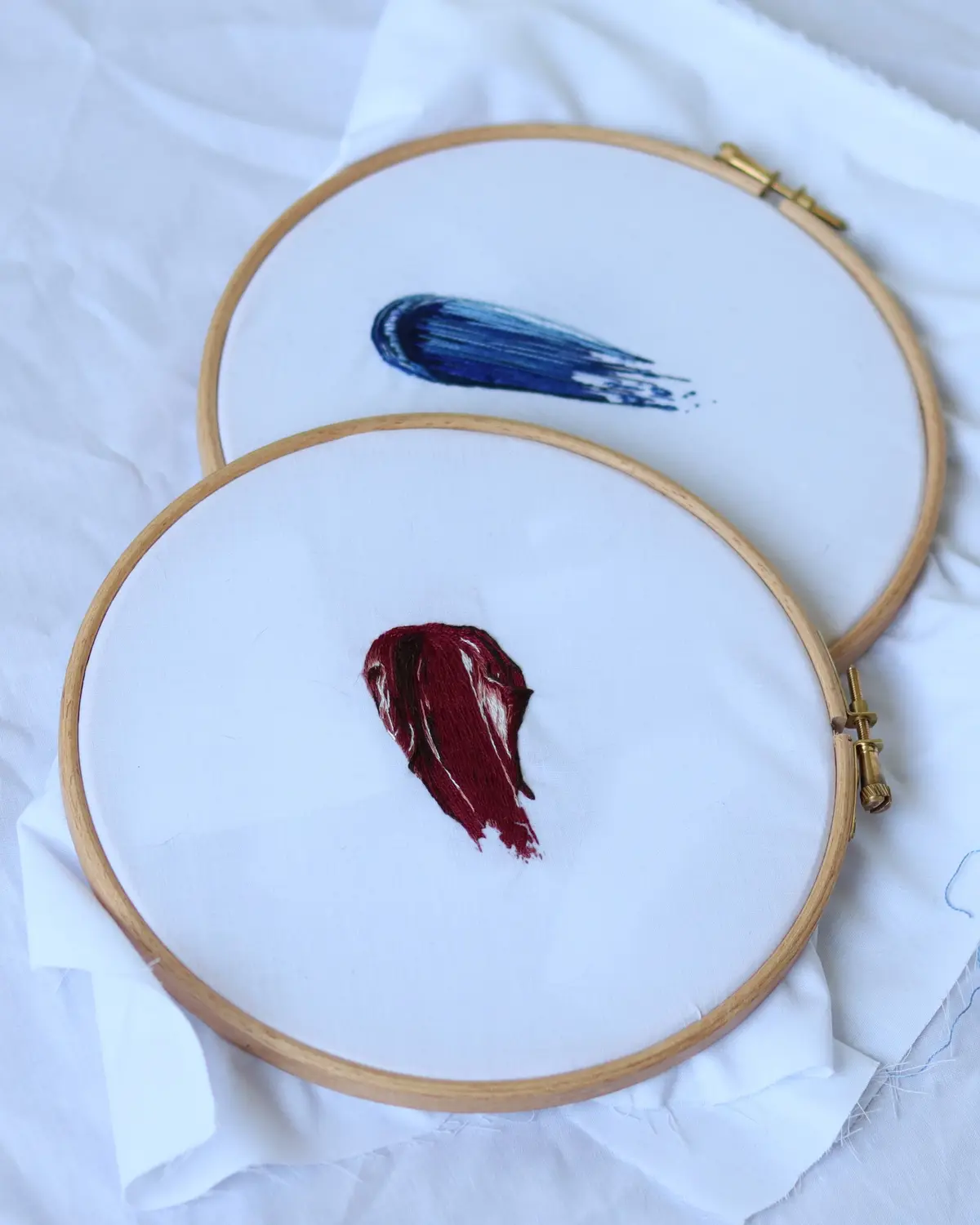 Two embroidery hoops on white fabric show abstract stitched shapes: a blue streak above a red, heart-shaped stain. The…