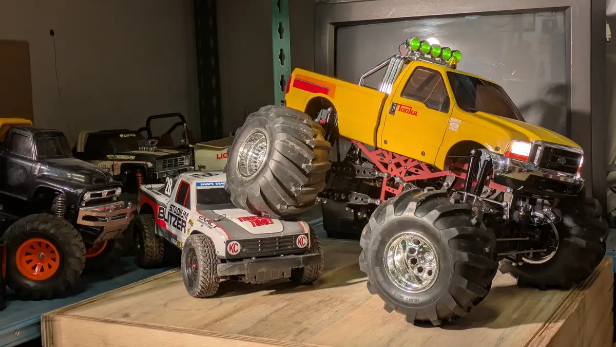 A large yellow Tonka monster truck with oversized black tires and green roof lights is shown crushing a smaller white and…