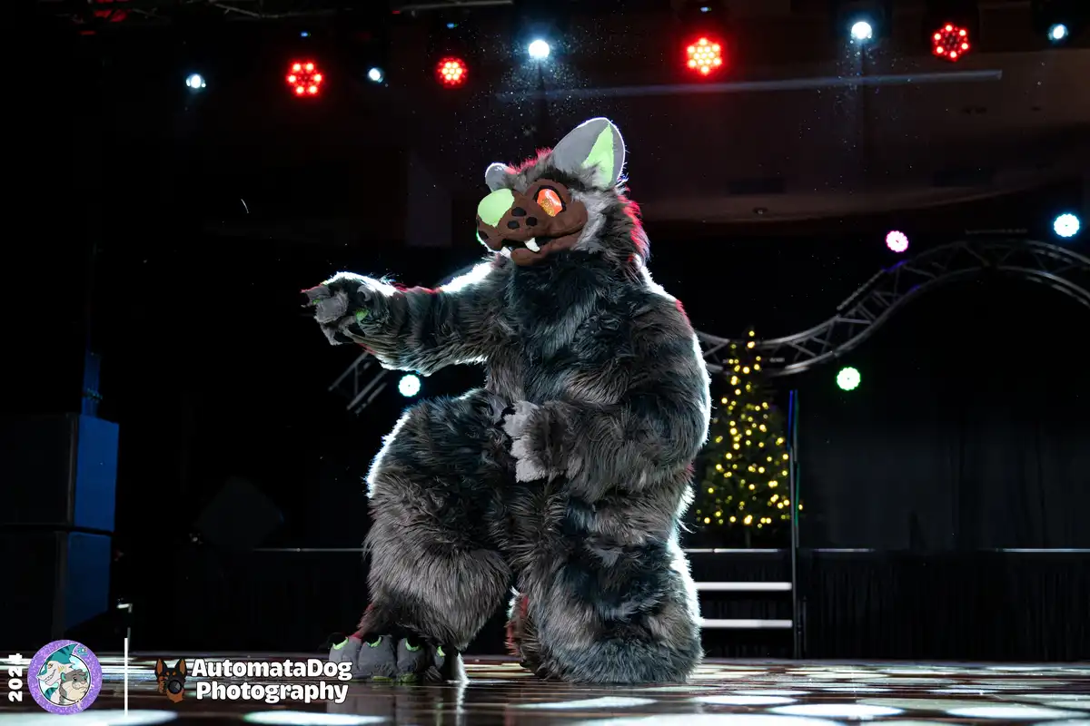 A person in a detailed, grey-furred, wolf-like fursuit with glowing orange eyes and green accents stands on a stage under…