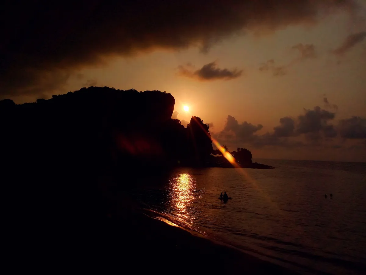 A serene sunset at a beach with the sun low on the horizon, casting a golden glow over the water and silhouetting cliffs and…