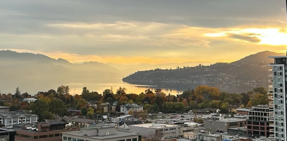 A scenic photo of a cityscape at sunset, featuring a large body of water, distant mountains, and autumn-colored trees, with…
