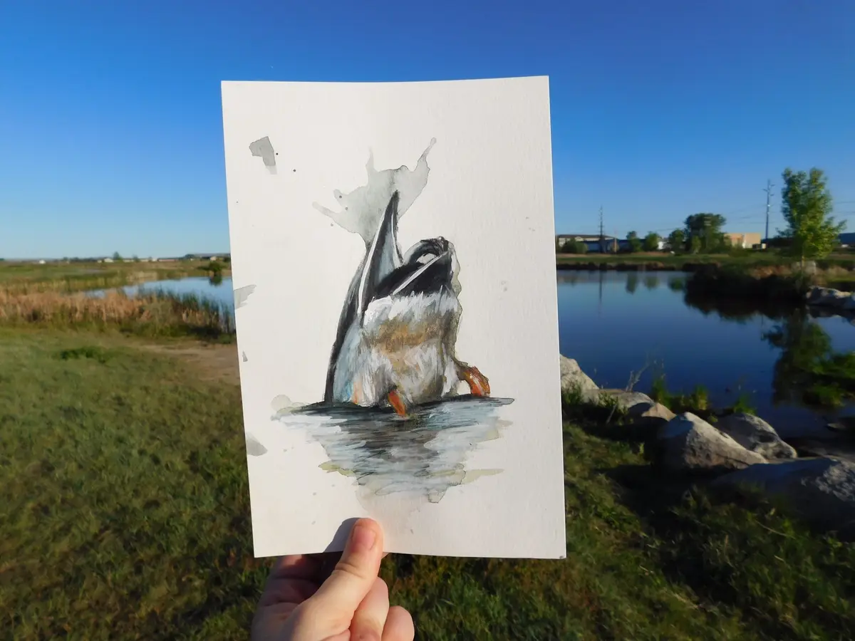 A hand holds up a watercolor painting of a duck splashing in water, positioned in front of a real pond under a clear blue…