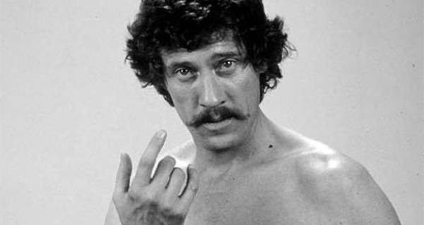 a black and white photo of a shirtless man with curly hair and a mustache, making a hand gesture with his fingers extended…