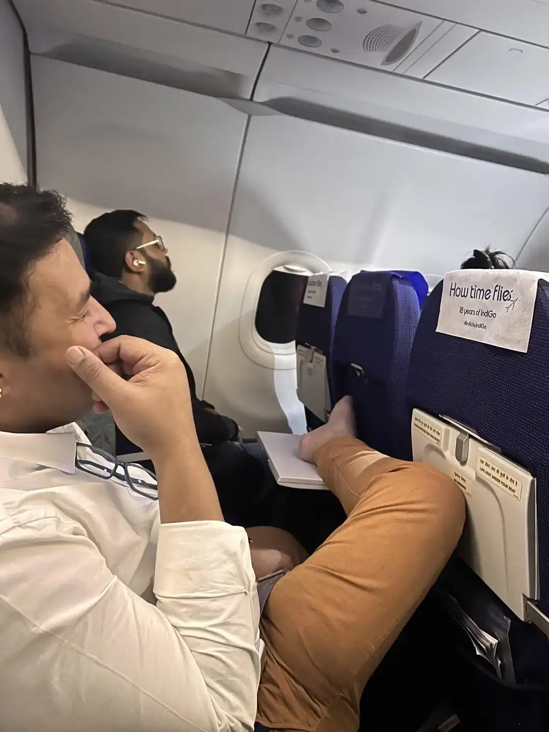 A man in a white shirt and brown pants sits in an airplane seat with his legs up on the tray table, looking thoughtful while…