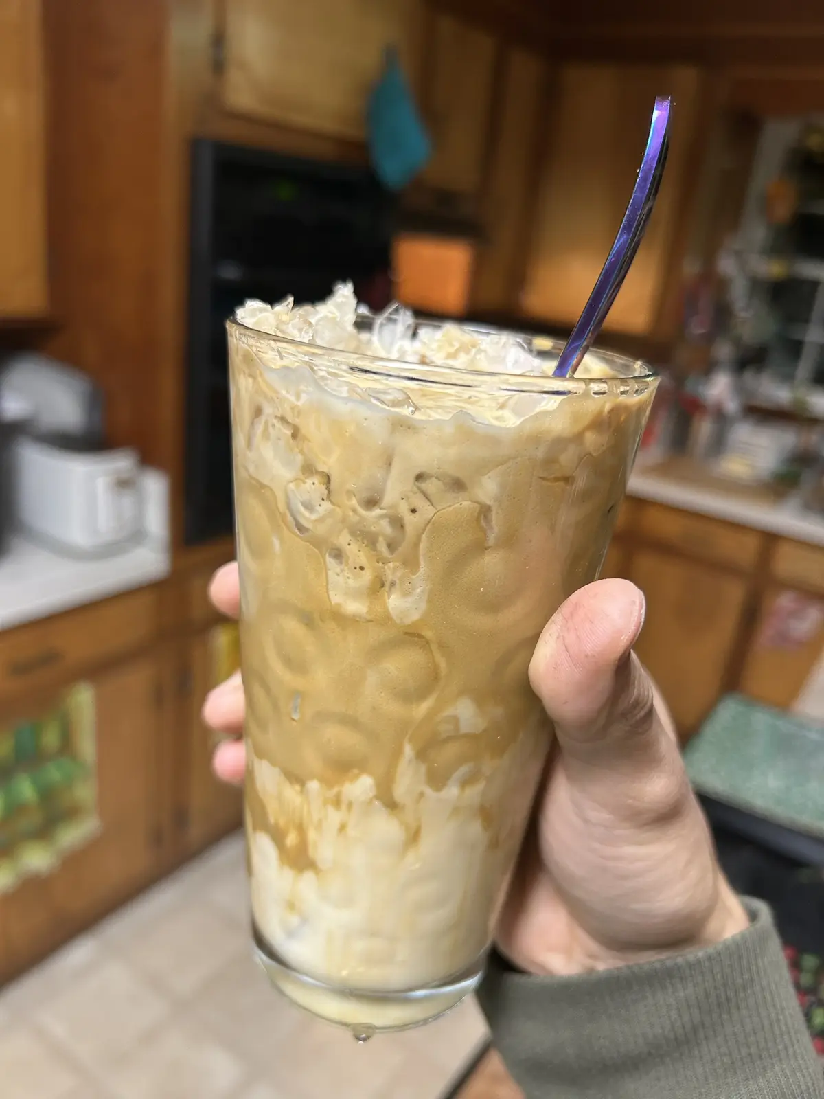 A hand holds a tall glass of iced coffee with ice cubes, cream, and a purple spoon, set against a blurry kitchen background…