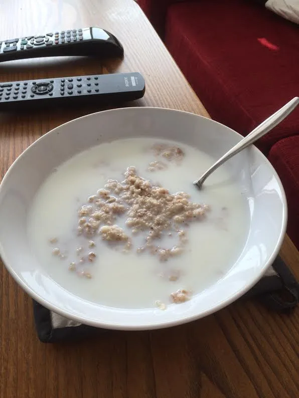 A white bowl of oatmeal with milk and a spoon sits on a wooden table next to two black remote controls, with a red couch in…