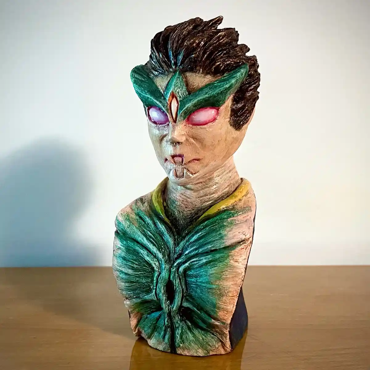 A handmade bust of a humanoid alien figure with glowing pink eyes, green facial markings, and brown hair, wearing a textured…