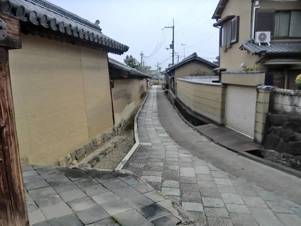 a narrow, winding alleyway in a residential area with traditional Japanese-style tiled roofs on buildings on both sides…