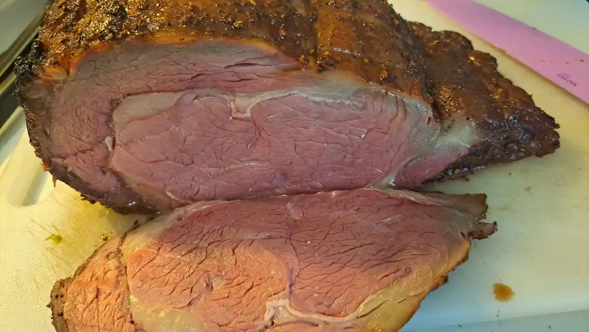 A close-up photo shows a large, sliced roast beef on a white cutting board, with one thick slice cut and resting in front…