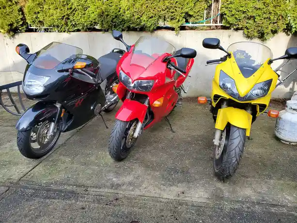 Three Honda sport motorcycles, black, red, and yellow, are parked side-by-side on a concrete surface in front of a concrete…