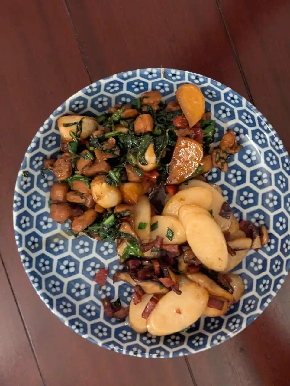 A top-down photo of a plate with two distinct food portions: one side has stir-fried chicken, greens, and garlic cloves; the…