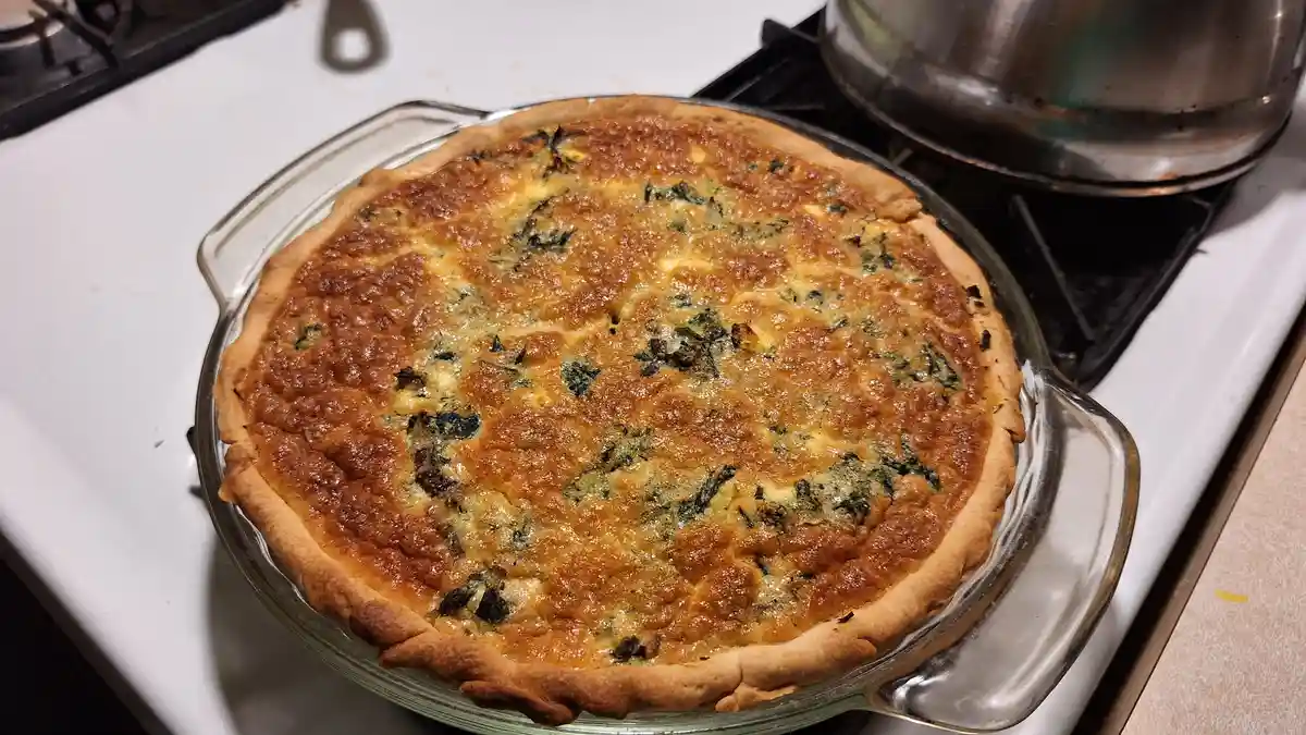 A golden-brown quiche with a fluted crust sits in a clear glass pie dish on a white stovetop, next to a metal pot. The text…