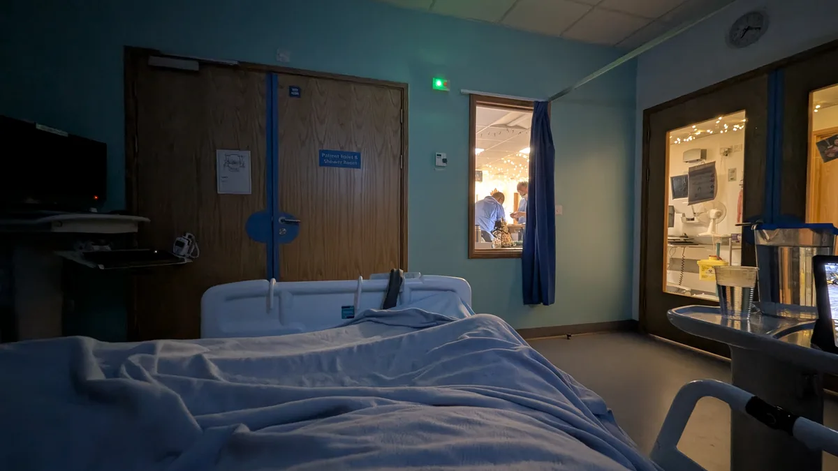 A dimly lit hospital room with a patient bed in the foreground, featuring blue walls and a wooden door labeled 'Patient…